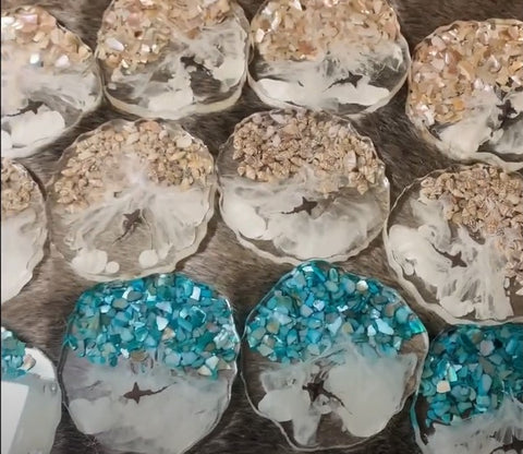 Several circular sea coasters made with epoxy resin, white pigment, shiny teal glass pieces and seashells. Shark silhouette in the middle.