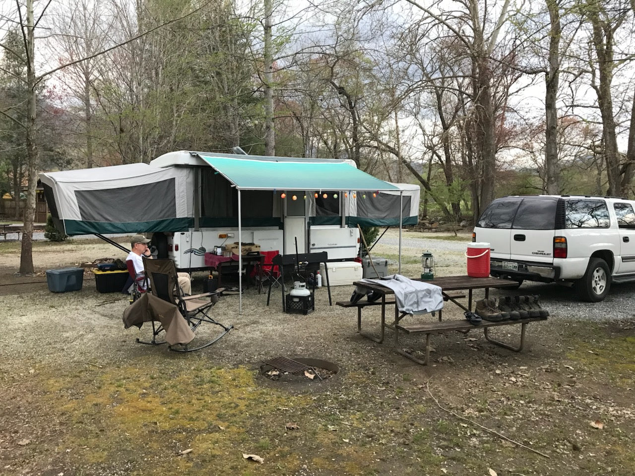 Finished pop up tent trailer set up in campground with picnic table, man and car sitting in front of it.