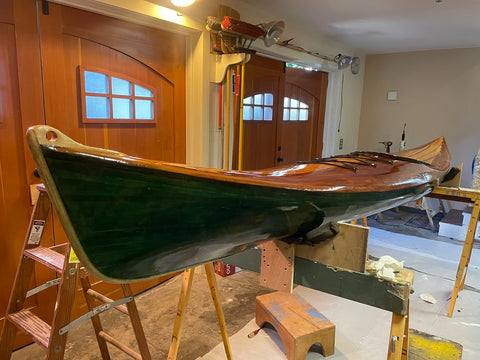 Side view of green and dark brown wood kayak that has a shiny resin finish. Sitting on a stand in a workshop.
