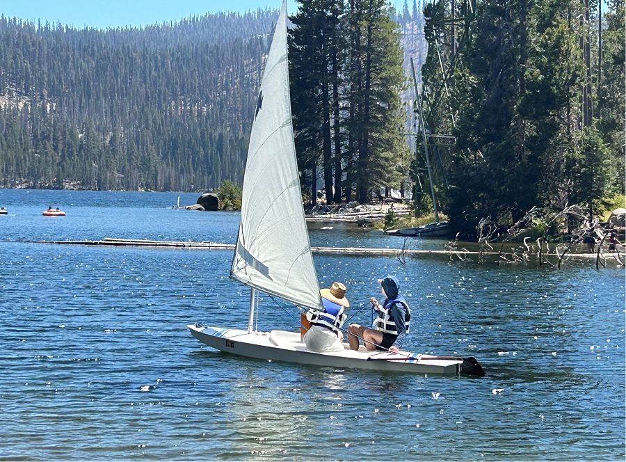 Newly resurfaced sunfish sailboat on the water with two people sailing on it in the mountains.